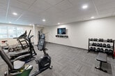 Located at 225 Sandwich St N in Amherstburg, Ontario, this fitness facility offers a well-appointed gym space designed to support a comprehensive workout routine. The room features a drop-panel ceiling with recessed lighting that evenly illuminates the entire area, creating a bright and functional environment. Durable, patterned carpet tile flooring covers the floor throughout, providing both comfort and practicality underfoot.

The gym is equipped with a varied selection of cardio machines, including a treadmill, stationary bikes, an elliptical trainer, and an upright cycle, catering to a range of cardiovascular fitness needs. A multi-function cable and weight machine is positioned along one wall for strength training exercises. A tiered dumbbell rack stocked with free weights, accompanied by a flat weight bench, provides additional options for resistance training.

A flat-screen TV is wall-mounted above a floating media shelf, allowing users to watch content during their workouts. Natural light enters through windows fitted with vertical blinds, contributing to an open and energizing atmosphere. A wall-mounted air conditioning unit ensures climate control throughout the space. The layout is clean and efficient, with equipment arranged to maximize usable floor space while maintaining clear pathways for safe movement throughout the gym.