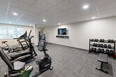 Located at 225 Sandwich St N in Amherstburg, Ontario, this fitness facility offers a well-appointed gym space designed to support a comprehensive workout routine. The room features a drop-panel ceiling with recessed lighting that evenly illuminates the entire area, creating a bright and functional environment. Durable, patterned carpet tile flooring covers the floor throughout, providing both comfort and practicality underfoot.

The gym is equipped with a varied selection of cardio machines, including a treadmill, stationary bikes, an elliptical trainer, and an upright cycle, catering to a range of cardiovascular fitness needs. A multi-function cable and weight machine is positioned along one wall for strength training exercises. A tiered dumbbell rack stocked with free weights, accompanied by a flat weight bench, provides additional options for resistance training.

A flat-screen TV is wall-mounted above a floating media shelf, allowing users to watch content during their workouts. Natural light enters through windows fitted with vertical blinds, contributing to an open and energizing atmosphere. A wall-mounted air conditioning unit ensures climate control throughout the space. The layout is clean and efficient, with equipment arranged to maximize usable floor space while maintaining clear pathways for safe movement throughout the gym.