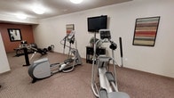 This fitness room provides a dedicated space for cardiovascular exercise, featuring multiple elliptical trainers and additional cardio equipment arranged throughout the carpeted area. The room is finished with neutral-toned walls and a standard drop ceiling with recessed lighting, creating a clean and functional atmosphere. A wall-mounted flat-screen television is positioned on a freestanding stand, allowing residents to stay entertained during workouts. A dark-wood storage console sits beneath the TV, accompanied by a small decorative plant that adds a touch of warmth to the space. Framed artwork on the walls contributes to a welcoming ambiance. The carpeted flooring provides a softer surface underfoot, and the overall layout allows for comfortable movement between machines. The room appears well-maintained and thoughtfully organized, making it a practical amenity for those seeking a convenient on-site fitness option.