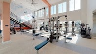 Located in Fort Worth, TX, this two-story fitness center is a well-appointed facility designed to accommodate a comprehensive range of workout needs. The main floor is characterized by soaring double-height ceilings flooded with natural light from an expansive wall of large windows, creating a bright and energizing environment. Overhead, large ceiling fans maintain airflow throughout the space. The main floor is equipped with an extensive lineup of cardio machines — including treadmills, ellipticals, and StairMaster units — alongside a full array of cable machines, weight benches, and a dedicated dumbbell rack station. Mounted flat-screen televisions are positioned throughout for viewing convenience. A standout design feature is the vivid, large-scale mural depicting iconic Fort Worth and Texas landmarks, adding a strong sense of local identity to the space. A metal staircase with industrial-style railings connects the main floor to the upper mezzanine level, which offers additional equipment and overlooks the main workout area below. A separate enclosed studio room provides a dedicated space for functional training, featuring floor-to-ceiling mirrors, TRX suspension systems, kettlebells, medicine balls, and wall-mounted screens. The overall aesthetic is modern and clean, with warm coral accent beams complementing the neutral flooring and white walls.