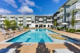 Located at 83 Ernest W Barrett Pkwy NW in Marietta, Georgia, this community amenity space combines contemporary design with resort-style comfort. The centerpiece of the outdoor area is a large, sparkling swimming pool framed by an expansive sun deck furnished with lounge chairs, dining tables, and shaded umbrella seating. An outdoor grilling and lounge station provides an ideal setting for social gatherings, all set against a lush, tree-lined backdrop.

Inside, the clubhouse showcases a refined interior defined by striking herringbone-patterned hardwood flooring that flows throughout the open layout. Structural columns finished in rich dark wood add architectural character, while recessed lighting and a series of elegant pendant fixtures—including distinctive woven cage-style lanterns—illuminate the space warmly. A dramatic double-height atrium allows abundant natural light to pour in through floor-to-ceiling windows, offering treetop views of the surrounding greenery.

A gallery-style corridor features a curated art display mounted on sage green paneled walls, including large-format photography that adds a thoughtful, artistic touch. Open-railed staircases with black iron balusters connect the levels, reinforcing the space's modern aesthetic. Throughout, the design balances clean white walls, natural materials, and carefully selected furnishings to create an inviting and well-appointed community environment.