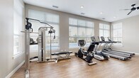 This fitness room is a thoughtfully designed exercise space that offers a comprehensive workout environment within a compact, efficiently utilized floor plan. The room features warm-toned hardwood-style flooring throughout, neutral gray walls, and a clean white ceiling fitted with recessed lighting and a ceiling fan to maintain comfortable airflow during workouts.

Large windows with horizontal blinds line the walls, flooding the space with abundant natural light and offering views of the surrounding outdoor scenery. The cardio section is well-appointed with multiple True-brand treadmills, an elliptical machine, and a recumbent stationary bike, all positioned to take advantage of the natural light from the windows.

Strength training is well-represented with a multi-function cable machine and a dedicated weight bench. A full rack of dumbbells is neatly organized alongside a large wall mirror, providing functionality for free-weight exercises. The space also includes a medicine ball rack with a range of weighted balls and stability/exercise balls, along with an interlocking foam mat area designated for stretching or floor exercises.

A water cooler station adds a practical amenity, and framed artwork on the wall contributes a polished, welcoming atmosphere. The layout maximizes usability while maintaining open floor space for versatile workouts.