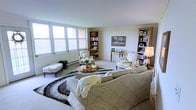 This single-floor space centers around a generously sized living room that combines classic elegance with functional design. Large multi-pane windows along one wall flood the room with natural light, creating a bright and airy atmosphere. The neutral color palette, featuring soft whites and warm beiges on the walls and ceiling, enhances the sense of openness throughout.

The living area is thoughtfully arranged with a plush, oversized sofa accented by a cozy throw blanket, complemented by two upholstered armchairs in a traditional French provincial style. A round wooden coffee table sits atop a bold black-and-white patterned area rug, adding visual interest to the seating arrangement. A tufted ottoman provides additional seating near the window.

Storage and display are well addressed through a combination of freestanding and built-in bookshelves positioned around the room, offering ample space for books, decorative items, and personal mementos. A small writing desk with a mirror occupies one corner, adding a refined, functional touch. Wall art and a table lamp contribute warmth, rounding out the room's classic, welcoming ambiance.