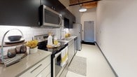 This single-floor space showcases a distinctive blend of modern and industrial design elements throughout. The most striking architectural feature is the natural wood tongue-and-groove ceiling with exposed timber beams, which adds warmth and character to the otherwise clean, contemporary interior. Smooth, light-toned flooring runs consistently throughout the unit, complemented by crisp white walls and dark baseboard trim that creates a sharp visual contrast.

The open living area is generously proportioned, featuring recessed lighting integrated into the wood ceiling, a wall-mounted PTAC heating and cooling unit, and a window with a roller shade that allows for controlled natural light and views to the outdoors. Multiple electrical outlets are positioned throughout the space, offering flexibility for various layouts and uses.

The kitchen is well-appointed with a full suite of stainless steel appliances, including a range, microwave, dishwasher, and refrigerator. Dark upper cabinetry contrasts with lighter lower cabinets, and a white subway tile backsplash adds texture. A pendant light fixture overhead enhances the functional workspace.

A hallway leads to a full bathroom with a vanity, mirror, and toilet, alongside a utility closet and an electrical panel. The overall ambiance is clean, functional, and modern.