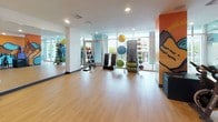 This fitness studio is a dynamic, single-floor space designed to support a variety of workout routines. The layout is divided into distinct functional zones, each thoughtfully outfitted for different exercise disciplines. The open workout area features smooth hardwood-style flooring, recessed lighting, and floor-to-ceiling windows that allow generous natural light and outdoor views. Racks of medicine balls and stability balls in multiple sizes line the walls, alongside step platforms and other functional training equipment. A large wall-mounted mirror enhances the sense of space in this zone. The adjoining cycling and yoga studio is a standout feature, showcasing a bold, large-scale mural depicting a mountainous landscape in vivid orange, blue, and black tones. A row of spin bikes faces the mural, creating an energetic atmosphere for cycling sessions. Glass partition walls etched with "YOGA" define the studio's boundaries while maintaining visual connectivity throughout the space. A wall-mounted flat-screen television adds a modern touch. Together, the areas create a cohesive, energetic fitness environment that balances open-concept training with dedicated studio functionality.