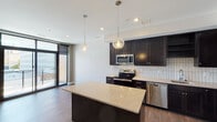 This contemporary single-floor residence showcases a thoughtfully designed open-concept layout that seamlessly connects the kitchen and living areas. The kitchen is a standout feature, equipped with rich dark cabinetry, a classic white subway tile backsplash, stainless steel appliances including a range, microwave, dishwasher, and refrigerator, and a generous center island topped with light stone countertops. Globe pendant lights hang above the island, adding a stylish focal point to the space. Wood-look flooring runs throughout the main living areas, contributing warmth and continuity.

The bedrooms offer plush carpeting underfoot and benefit from expansive floor-to-ceiling windows that frame urban city views, flooding the rooms with natural light. One bedroom opens directly to a private outdoor balcony through a sleek glass door, providing convenient outdoor access. The bathroom is finished with large-format tile flooring, a dark vanity with a stone countertop, a framed mirror, and clean, neutral walls that create a calm, functional environment.

Throughout the residence, a neutral color palette of soft whites and warm grays creates a clean, modern aesthetic. Recessed lighting complements the natural light, ensuring a bright and welcoming atmosphere across all rooms. The overall design is both functional and contemporary, well-suited for urban living.