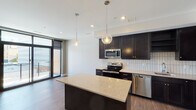 This contemporary single-floor residence showcases a thoughtfully designed open-concept layout that seamlessly connects the kitchen and living areas. The kitchen is a standout feature, equipped with rich dark cabinetry, a classic white subway tile backsplash, stainless steel appliances including a range, microwave, dishwasher, and refrigerator, and a generous center island topped with light stone countertops. Globe pendant lights hang above the island, adding a stylish focal point to the space. Wood-look flooring runs throughout the main living areas, contributing warmth and continuity.

The bedrooms offer plush carpeting underfoot and benefit from expansive floor-to-ceiling windows that frame urban city views, flooding the rooms with natural light. One bedroom opens directly to a private outdoor balcony through a sleek glass door, providing convenient outdoor access. The bathroom is finished with large-format tile flooring, a dark vanity with a stone countertop, a framed mirror, and clean, neutral walls that create a calm, functional environment.

Throughout the residence, a neutral color palette of soft whites and warm grays creates a clean, modern aesthetic. Recessed lighting complements the natural light, ensuring a bright and welcoming atmosphere across all rooms. The overall design is both functional and contemporary, well-suited for urban living.