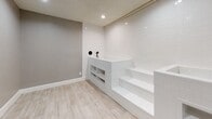 This single-level space is thoughtfully designed around a dedicated wash or grooming station, making it a highly functional and purpose-built area. The centerpiece of the room is an elevated, white subway-tiled platform accessible via built-in steps, providing convenient access to the washing area. Matte black wall-mounted fixtures, including a handheld sprayer, add a modern, contrasting detail against the crisp white tilework. Integrated open shelving is built directly into the platform structure, offering practical storage for grooming supplies or accessories within easy reach.

The flooring throughout consists of light wood-look planks that lend warmth and a clean aesthetic to the space. Walls are finished in a soft gray tone with a textured surface, complemented by bright white baseboards that create a sharp, polished contrast. Recessed lighting in the textured ceiling provides even, functional illumination across the room. A panel door with a classic raised design provides access to the space, while a secondary wall with a glimpse of natural light hints at an adjoining area. The overall design balances practicality with a clean, contemporary style.