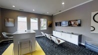 This single-floor space presents a clean, contemporary design that blends functionality with modern aesthetics. The main area is anchored by a crisp white reception desk, offering a polished first impression for visitors. A row of minimalist white chairs with chrome frames lines the room, providing comfortable seating alongside a low, white storage credenza that spans the adjacent wall. Above the credenza, multiple flat-screen displays are mounted, adding a dynamic, media-ready element to the space. The walls are finished in a sophisticated warm gray tone, complemented by bright white crown molding and a white ceiling fitted with recessed lighting that ensures even, well-distributed illumination throughout. A bold yellow accent on the floor near the reception desk introduces a vibrant pop of color, contrasting tastefully against the patterned dark blue-gray carpeting that covers the majority of the floor. A series of windows along one wall invites natural light into the space, offering views of lush greenery outside. Framed artwork on the walls adds a curated, professional touch. Overall, the space conveys a welcoming yet efficient atmosphere well-suited for client-facing or collaborative professional use.