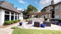This single-story space encompasses two distinct areas: a thoughtfully designed interior mailroom and an inviting outdoor communal patio. The mailroom features soaring ceilings painted in a cool blue-gray tone, creating an airy and open atmosphere. Light-colored tile flooring reflects natural light streaming through large windows, enhancing the sense of spaciousness. A central island unit finished in warm bronze-toned cabinetry houses an extensive array of individual mailboxes, with additional wall-mounted units illuminated by integrated LED strip lighting. Framed geometric artwork adorns the walls, adding a contemporary decorative element to the functional space.

The outdoor area showcases a generously sized flagstone patio anchored by a wooden pergola structure that shelters a built-in outdoor kitchen equipped with multiple grills and stone countertops. Dark wicker lounge seating is arranged throughout the patio, complemented by vibrant cobalt blue planters. Mature trees provide natural shade and a lush backdrop, while manicured shrubs and lawn areas frame the space. The surrounding buildings feature classic stone and brick architecture with arched colonnades, contributing to an elegant and cohesive communal environment.
