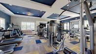 Located at 1500 German School Road in Richmond, VA, this fitness center offers a well-appointed, single-floor exercise room designed to support a comprehensive workout experience. The space is defined by its striking navy blue accent walls and matching tray ceilings trimmed in white, creating a bold yet polished aesthetic. Recessed lighting throughout ensures bright, even illumination across the entire floor.

The exercise room is generously sized, providing ample space to accommodate a wide range of fitness equipment without feeling overcrowded. Cardio equipment includes treadmills, ellipticals, and stationary bikes, several of which feature interactive display screens. The strength training area is equally well-equipped, with cable machines, a power rack, a weight bench, barbells, and a full dumbbell rack. A punching bag and additional functional fitness accessories round out the offerings.

Large, arched windows with white plantation shutters allow natural light to filter into the space, enhancing the open and energizing atmosphere. Full-length mirrors are strategically placed along the walls, visually expanding the room while providing practical utility for form checks during workouts. The floor features a patterned carpet tile in neutral, navy, and gold tones, adding texture and visual interest while providing cushioned support underfoot. Overall, this fitness center is a thoroughly equipped and attractively designed space.