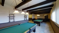 Located at 2311 West 16th Street in Greeley, Colorado, this single-floor recreational space is thoughtfully designed for leisure and social entertainment. The room is anchored by multiple full-sized billiard tables finished in rich dark wood with green felt surfaces, each illuminated by elegant pendant lighting fixtures suspended from above. The ceiling features a series of bold, dark exposed beams that run the length of the space, adding a rustic yet refined architectural character. Light-toned tile flooring throughout provides a clean, durable foundation that complements the darker tones of the furnishings. Along the walls, dedicated cue racks keep equipment neatly organized and accessible. Toward the rear of the space, a lounge area is furnished with seating, creating a comfortable area for spectators or casual conversation. Additional pendant lighting is distributed throughout, ensuring the entire area is well-lit. The overall ambiance strikes a balance between a classic game room aesthetic and a welcoming communal environment, making it well-suited for both competitive play and relaxed social interaction.