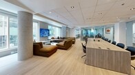 Located at 1009 Rue Bleury in the heart of Montréal, this thoughtfully designed lounge and conference space offers a versatile, professional environment across a generous single-floor layout. The space seamlessly blends collaborative open areas with dedicated private meeting rooms, catering to a wide range of professional needs.

The open lounge area is anchored by a large light wood conference table surrounded by modern seating, flowing naturally into a comfortable lounge zone furnished with plush sectional sofas and a wall-mounted flat-screen display. Large windows allow abundant natural light to fill the space, framing views of the surrounding urban streetscape and lending an airy, welcoming ambiance.

Floor-to-ceiling glass partitions define the enclosed meeting rooms without sacrificing visual openness, maintaining a bright and connected atmosphere throughout. One standout room features a bold design statement: a sculptural oval pendant light fixture overhead, a large presentation screen on a dark accent wall, lush potted greenery, and vibrant orange and navy seating arranged around a sleek white conference table.

A kitchenette and bar area with pendant lighting and counter-height seating provides a functional social hub within the space. Light hardwood flooring runs throughout, complementing the clean white ceilings with recessed lighting. Together, these elements create a polished, contemporary professional environment suited for meetings, events, and everyday collaboration.
