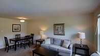 This single-floor residence offers a welcoming and efficiently arranged living environment. The combined living and dining area serves as the heart of the home, creating an open, cohesive space ideal for both relaxing and entertaining. The living area is anchored by a plush gray sofa adorned with decorative throw pillows, complemented by a dark-finished coffee table and side tables that add a touch of contrast. Warm ambient lighting from table lamps enhances the soft, neutral tones of the walls, creating a cozy atmosphere throughout. A framed botanical wall print adds a subtle decorative accent, contributing to the space's understated charm. Adjacent to the living area, a dining set with dark wood chairs provides a functional and intimate spot for meals. Soft wall-to-wall carpeting runs throughout the space, adding warmth and comfort underfoot. The smooth, flush ceiling with a centrally placed light fixture keeps the design clean and uncluttered. Overall, the space presents a harmonious blend of neutral tones, practical layout, and comfortable furnishings, making it a well-appointed and livable home.