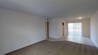 This single-floor residence offers a clean, functional layout designed for comfortable everyday living. The main living area is finished with wall-to-wall carpeting, providing a warm and inviting atmosphere, while the adjoining transition spaces feature light-toned ceramic tile flooring that extends toward the kitchen and other areas of the home. Crisp white walls throughout the space contribute to a bright, airy feel, complemented by a textured ceiling that adds subtle dimension overhead. A sliding glass door dressed with vertical blinds allows natural light to filter into the living area while offering direct access to an outdoor space, enhancing the sense of openness. A partial wall divides the living area from the adjoining tiled zone, creating a natural flow between spaces without fully closing them off. A doorway visible from the main living area leads to an interior room, suggesting a well-connected floor plan. The kitchen, partially visible in the background, appears to include standard appliances. Overall, the space presents a straightforward, well-lit interior with a neutral palette and practical finishes suited to a wide range of furnishing styles.