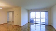 This single-floor space offers a clean, open layout defined by its polished hardwood flooring that reflects natural light throughout the interior. The walls are finished in a crisp, neutral white, creating a bright and airy atmosphere that feels welcoming and versatile. A standout feature of the main living area is the expansive sliding glass door, which spans nearly the full width of the exterior wall and opens onto a balcony. This large glazed opening floods the space with abundant natural light while providing views of the surrounding neighborhood. The balcony itself features a railing and is visible beyond the glass, offering an outdoor extension of the living area. A thermostat and multiple electrical outlets are positioned along the walls, indicating practical utility throughout. An open doorway visible along one wall connects to an adjoining room or hallway, suggesting a functional flow between spaces. The overall ambiance is minimal and move-in ready, with a neutral palette and quality finishes that provide a solid foundation for a variety of interior arrangements and design styles.
