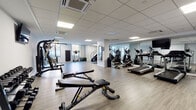 This fitness center at 720 Bronx River Rd in Yonkers, NY offers a spacious, single-level layout designed to accommodate a full range of workout activities. The facility features light wood-style flooring throughout, a drop ceiling with recessed panel lighting, and a climate control system to maintain a comfortable environment during exercise.

The cardio area is well-stocked with treadmills, elliptical machines, and a stair climber, positioned alongside large floor-to-ceiling mirrors that visually expand the space and allow users to monitor their form. Natural light filters in through windows on one side of the room, contributing to a bright and open atmosphere.

The strength training section includes a multi-station cable machine, dedicated weight benches, adjustable incline benches, and a dumbbell rack with a complete set of free weights. Wall-mounted televisions are positioned throughout the space for entertainment during workouts.

An additional area within the facility includes a medicine ball rack with an assortment of weighted balls, a treadmill, and a water fountain station with both a standard drinking fountain and a bottle-filling spout, set against white subway tile accents. The overall design is clean, functional, and modern, providing a well-rounded environment suited to a variety of fitness routines.