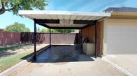 This Car Care Center is a single-level, open-air car washing facility designed to provide residents with a convenient and functional vehicle maintenance space. The structure features a sturdy metal roof supported by dark steel posts, offering overhead coverage while remaining open on multiple sides to allow for natural ventilation and airflow during use.

The wash bay is finished with a sealed concrete floor and includes a central floor drain for efficient water runoff, keeping the area clean and manageable during and after use. A water hose connection is mounted and accessible on one side of the bay, making vehicle rinsing and washing straightforward. An adjacent brick enclosure on the side of the structure houses utility equipment, adding to the overall infrastructure of the space.

The facility is enclosed on one side by a tall wooden privacy fence, while the opposite side opens toward a landscaped community streetscape lined with mature trees and green lawns. A sign above the entrance clearly identifies the space as the "Car Care Center." The combination of durable construction materials, practical drainage, and an open design makes this a well-equipped and user-friendly amenity within the community.