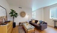 This single-floor, one-bedroom, one-bathroom residence offers a well-organized layout that makes efficient use of its compact footprint. Light hardwood flooring flows throughout, unifying the living, kitchen, and hallway spaces in a warm, cohesive aesthetic.

The living room features clean white walls, a ceiling-mounted HVAC vent, and a window that draws in natural light. The space accommodates a comfortable seating arrangement alongside a glass-top coffee table and a woven wood dresser, with decorative touches like a sunburst wall accent adding character.

The kitchen is outfitted with flat-front light wood cabinetry, a white penny-tile backsplash, and a full suite of stainless steel appliances including a refrigerator, range, microwave, and dishwasher. A large window above the counter provides ample natural light, and under-cabinet lighting enhances the workspace.

The bedroom is a calm, well-proportioned retreat featuring a window with exterior views and a wide double sliding-door closet with built-in shelving and hanging space, offering practical storage.

The bathroom presents a clean, modern design with large-format gray tile flooring, a floating wood vanity with a white countertop, an illuminated mirror, and crisp white walls that keep the space feeling bright and fresh.