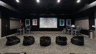 This 680 square foot living room has been transformed into a state-of-the-art home theater, offering a cinematic experience within a residential setting. The space features a sleek, modern design with dark walls and ceiling, creating an ideal environment for movie viewing. A large projection screen serves as the focal point, complemented by a high-quality projector mounted on the ceiling.

The seating arrangement is tiered, mimicking a commercial theater layout. It includes plush, black leather recliners in the front row, providing premium comfort for viewers. Additional seating is available in the form of oversized bean bags, offering a more casual and versatile seating option.

The room's aesthetics are enhanced by colorful, abstract artworks adorning the walls, adding visual interest and contrast to the predominantly dark space. Ambient lighting is strategically placed to create the right atmosphere without interfering with the viewing experience.

For added convenience, the space includes a small bar area with high stools, perfect for refreshments. The flooring consists of patterned carpeting, which aids in sound absorption for optimal acoustics. An exit door with an illuminated sign ensures safety compliance.

This thoughtfully designed home theater maximizes the available space, creating a multi-functional entertainment area that combines comfort, style, and cutting-edge technology.
