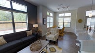 This single-floor space presents a thoughtfully designed open-concept layout that seamlessly connects the living and dining areas into one cohesive, functional environment. The living area is anchored by a sleek dark sofa and complementary plaid accent chairs, set atop hardwood-style flooring that runs throughout the space, lending warmth and visual continuity. A bold dark accent wall adds depth and contrast against the light neutral tones of the surrounding walls, creating a striking focal point.

The space benefits from an abundance of natural light, thanks to multiple large windows positioned along the walls, including elevated transom-style windows that draw the eye upward and emphasize the impressive ceiling height. These windows offer views of the surrounding greenery, further enhancing the airy, open atmosphere.

The dining area is defined by a contemporary table paired with bold yellow chairs, introducing a vibrant pop of color that energizes the space. Track lighting on the ceiling provides targeted illumination, complementing the natural light throughout the day. The overall ambiance is modern yet inviting, with a carefully curated palette of charcoal, yellow, and neutral tones that balances style with comfort, making this a versatile and welcoming space.