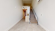 Located at 974 Front Ave NW in Grand Rapids, Michigan, this two-floor loft-style unit offers a distinctive industrial-modern aesthetic spread across two levels. The main floor serves as the primary living area, featuring polished tile flooring with a marble-like finish, crisp white walls, and soaring ceilings with exposed painted beams that lend a raw, warehouse-inspired character. A built-in kitchen peninsula with a light wood finish and integrated sink anchors the space, while track lighting mounted along the ceiling provides focused illumination throughout. A carpeted staircase with a sleek black metal handrail connects the main floor to the upper mezzanine level. The mezzanine, finished with carpet underfoot and the same striking exposed beam ceiling, overlooks the main level and is bathed in natural light from large industrial-style grid windows that frame views of the surrounding neighborhood. The bathroom is thoughtfully appointed with warm wood cabinetry featuring matte black hardware, a white countertop vanity, and polished tile flooring that complements the main living area. Stainless grab bars add a practical touch. A hallway and two staircase areas complete the layout, contributing to the efficient and well-connected flow between floors. This unit combines urban industrial character with clean, contemporary finishes.