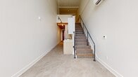 Located at 974 Front Ave NW in Grand Rapids, Michigan, this two-floor loft-style unit offers a distinctive industrial-modern aesthetic spread across two levels. The main floor serves as the primary living area, featuring polished tile flooring with a marble-like finish, crisp white walls, and soaring ceilings with exposed painted beams that lend a raw, warehouse-inspired character. A built-in kitchen peninsula with a light wood finish and integrated sink anchors the space, while track lighting mounted along the ceiling provides focused illumination throughout. A carpeted staircase with a sleek black metal handrail connects the main floor to the upper mezzanine level. The mezzanine, finished with carpet underfoot and the same striking exposed beam ceiling, overlooks the main level and is bathed in natural light from large industrial-style grid windows that frame views of the surrounding neighborhood. The bathroom is thoughtfully appointed with warm wood cabinetry featuring matte black hardware, a white countertop vanity, and polished tile flooring that complements the main living area. Stainless grab bars add a practical touch. A hallway and two staircase areas complete the layout, contributing to the efficient and well-connected flow between floors. This unit combines urban industrial character with clean, contemporary finishes.