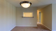 This single-floor residence offers a comfortable and practical layout designed for everyday living. The main living area is finished with plush wall-to-wall carpeting in a warm beige tone, creating a cozy and inviting atmosphere. A standout architectural detail is the pass-through window cut into the wall between the living and kitchen areas, complete with a small ledge — a classic design element that enhances connectivity between the two spaces while maintaining a degree of separation. The living area is illuminated by a centrally mounted bowl pendant light with a dark bronze finish, providing both functional and ambient lighting. The walls are painted in a soft, neutral beige that complements the carpeting and contributes to the overall warm feel of the space. White baseboards provide clean contrast against the walls and flooring. A hallway visible from the main living area leads further into the home, suggesting additional rooms beyond, including what appears to be a mirrored closet door. The space benefits from a neutral, move-in-ready aesthetic that offers a versatile backdrop for a variety of interior design styles.