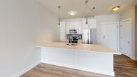 Located at 540 Revere Beach Blvd in Revere, MA, this one-bedroom, one-bathroom residence offers a well-designed layout within a modern multi-story building. The open-concept kitchen is a standout feature, showcasing white cabinetry, sleek quartz countertops, a peninsula with an undermount sink, stainless steel appliances including a refrigerator, microwave, and range, a subtle tile backsplash, and elegant pendant lighting. Wood-look flooring runs throughout the kitchen and living areas, lending warmth to the clean, contemporary aesthetic. The bedroom is a comfortable retreat with plush carpeting and a large window that fills the space with natural light, offering views of the building's landscaped courtyard or surrounding area. The bathroom is finished with light wood-look tile flooring, a glass-enclosed shower with subway tile surround, and crisp white fixtures. A private balcony overlooks the beautifully maintained central courtyard, surrounded by mature trees and communal outdoor spaces — ideal for relaxing and taking in the open-air setting. The building's exterior features a modern design with blue accent balconies and large windows throughout.