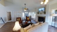 This single-floor residence offers a warm and inviting atmosphere centered around a thoughtfully designed open-concept living area. The main living space is anchored by a classic tiled fireplace with a clean white mantel, above which a wall-mounted television is positioned for comfortable viewing. A plush, neutral-toned sectional sofa pairs with rich, dark wood accent chairs accented with decorative gold-patterned throw pillows, creating an elegant yet cozy seating arrangement. A dark wood coffee table sits at the center of the space, completing the conversational layout.

Natural light pours in generously through a series of wide windows that line the walls, offering pleasant views of lush outdoor greenery and filling the interior with a bright, airy quality. Plantation shutters on select windows provide privacy and light control as needed. Ceiling fans with warm-toned fixtures enhance both the comfort and the classic character of the space.

The living area flows naturally into a dedicated dining space, visible through the open layout, where a dark wood dining set is bathed in natural light from the surrounding windows. Hardwood flooring ties the spaces together cohesively, adding warmth and continuity throughout. The overall ambiance is relaxed yet refined, making this home an ideal blend of comfort and classic style.