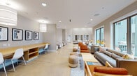 This contemporary community amenity space is thoughtfully designed across a single floor, offering a variety of functional zones that cater to diverse activities. Polished concrete floors run throughout, lending a clean, cohesive aesthetic that ties the space together seamlessly.

The main lounge area is generously proportioned, featuring large floor-to-ceiling windows that flood the interior with natural light and provide views of the surrounding outdoor courtyard. Comfortable seating arrangements — including plush sofas, leather accent chairs, and woven ottoman poufs — are grouped around low coffee tables, creating an inviting atmosphere for casual gatherings. Floor lamps add warm, ambient lighting throughout.

A dedicated work zone offers built-in wooden desks and sleek white chairs, ideal for focused tasks. Nearby, semi-private booth-style seating provides quieter spaces for individual use or small meetings. A separate dining area features a round table with modern white chairs alongside a wall-mounted counter workspace, accompanied by a tray ceiling detail and direct access to an outdoor balcony.

The media lounge stands out with its dramatic double-height ceiling, a bold herringbone wood accent wall, a wall-mounted flat-screen TV, and a colorful geometric area rug. A kitchenette with appliances rounds out this section. Motivational typographic artwork is displayed throughout, contributing to an energetic and modern community atmosphere.