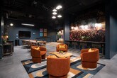 This single-floor space is designed with a distinct music-inspired aesthetic, combining moody, dark-toned interiors with vibrant artistic elements throughout. The main lounge area serves as the social centerpiece, featuring plush burnt-orange velvet armchairs accented with gold throw pillows, arranged around organic-shaped wood coffee tables atop a bold geometric area rug. Overhead, clustered pendant lights cast warm, focused illumination against the dark ceiling, which retains an exposed industrial quality.

A large-scale photographic mural of a live music festival—identified as the Capitol Hill Block Party—dominates one wall and anchors a bar counter lined with sleek gold-framed stools. Adjacent to this is a curated wall display of vintage loudspeakers, paired with a tan leather sofa and herringbone wood flooring, creating a distinct listening lounge nook. A flat-screen TV and communal dining table provide functional gathering options.

Semi-private booth areas enclosed by frosted glass partition doors offer intimate seating for smaller groups, each decorated with music-themed artwork such as a vintage microphone print and a dramatic portrait mural. A corner arcade game adds a playful touch. Throughout, the space balances industrial structure with warm, curated details, resulting in an immersive, entertainment-focused environment.