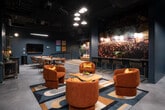 This single-floor space is designed with a distinct music-inspired aesthetic, combining moody, dark-toned interiors with vibrant artistic elements throughout. The main lounge area serves as the social centerpiece, featuring plush burnt-orange velvet armchairs accented with gold throw pillows, arranged around organic-shaped wood coffee tables atop a bold geometric area rug. Overhead, clustered pendant lights cast warm, focused illumination against the dark ceiling, which retains an exposed industrial quality.

A large-scale photographic mural of a live music festival—identified as the Capitol Hill Block Party—dominates one wall and anchors a bar counter lined with sleek gold-framed stools. Adjacent to this is a curated wall display of vintage loudspeakers, paired with a tan leather sofa and herringbone wood flooring, creating a distinct listening lounge nook. A flat-screen TV and communal dining table provide functional gathering options.

Semi-private booth areas enclosed by frosted glass partition doors offer intimate seating for smaller groups, each decorated with music-themed artwork such as a vintage microphone print and a dramatic portrait mural. A corner arcade game adds a playful touch. Throughout, the space balances industrial structure with warm, curated details, resulting in an immersive, entertainment-focused environment.