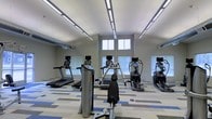 This fitness center is designed to support a well-rounded exercise routine in a clean, modern environment. The space features soaring ceilings that lend an open, airy feel, enhanced by rows of linear LED lighting that ensure consistent, bright illumination throughout. Large windows positioned near the upper portion of the walls allow natural light to filter in, contributing to an energizing atmosphere. The flooring consists of a patterned carpet tile in a mix of gray, blue, and white, adding a contemporary aesthetic while providing comfort underfoot. The equipment lineup includes multiple treadmills, elliptical cross-trainers, and a cable weight machine, all from commercial-grade brands, ensuring durability and performance. Mirrored wall sections along the perimeter expand the sense of space and allow users to monitor their form during workouts. The layout is thoughtfully organized, providing ample space between machines for safe and comfortable use. The neutral gray walls and clean design elements create a focused, distraction-free environment suited for fitness enthusiasts of all levels.