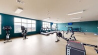 Located at 6834 Milwaukee St in Madison, Wisconsin, this fitness facility offers a spacious, single-floor exercise room designed to support a full range of workout routines. The room is finished with bold teal accent walls that create an energizing atmosphere, complemented by a neutral speckled floor that is both durable and easy to maintain. Flush-mounted linear lighting fixtures across the white ceiling ensure the space is consistently well-lit throughout.

Large windows along one wall allow ample natural light to filter in while providing views of the surrounding neighborhood, adding an open, airy feel to the environment. The cardio section is well-appointed with multiple treadmills, stationary bikes, an elliptical, and a rowing machine, offering diverse options for aerobic training. A dedicated strength area features a dumbbell rack with a full range of weights, a weight bench, and additional resistance equipment, supporting comprehensive strength training needs. Plyo boxes and functional fitness equipment round out the offerings for high-intensity training.

A wall-mounted flat-screen TV adds convenience for entertainment during workouts. The layout is thoughtfully organized, providing generous open floor space between equipment zones for safe, comfortable movement. An entry door with glass panels ensures easy access, completing this practical and well-designed fitness space.