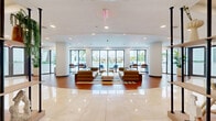 Located at 360 Granello Ave in Coral Gables, Florida, this single-floor community space offers a thoughtfully designed open-plan layout that seamlessly blends a fully equipped kitchen with a generous living area. The kitchen is appointed with rich warm-toned wood cabinetry, stainless steel appliances including a double wall oven and refrigerator, and a long quartz-topped island with bar seating — all illuminated by a row of sleek pendant lights. Adjacent dining tables provide additional seating for gatherings. The living area is divided into distinct lounge zones, each with its own character. One section features tan leather sofas, a glass coffee table, and a geometric-patterned area rug, all oriented toward floor-to-ceiling glass doors that open to views of a resort-style pool deck framed by palm trees. Another lounge area makes a bold design statement with a striking black-and-white op-art mural, built-in white shelving displaying curated décor and artwork, and contemporary soft seating arrangements. Throughout the space, warm wood-toned flooring, recessed lighting, and a bright white ceiling create a clean, modern ambiance. Structural cylindrical columns add an architectural element, contributing to the open, airy feel of this well-appointed communal space.