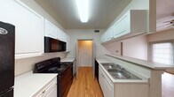 This single-floor residence offers a practical and well-organized layout designed for comfortable everyday living. The kitchen is a standout feature, designed in a classic galley configuration with white raised-panel cabinetry lining both sides. It is fully equipped with black appliances, including an electric range, over-the-range microwave, refrigerator, and dishwasher, all set against light countertops and warm hardwood-style flooring. A pass-through peninsula area connects the kitchen to the adjacent living space, which features vaulted ceilings that add a sense of openness and height. The living area and hallways continue the warm hardwood-style flooring, creating a cohesive flow throughout the main areas. The bedroom is generously sized, finished with plush carpeting and a ceiling fan for year-round comfort, and includes closet access. The bathroom is clean and functional, featuring a white vanity with ample counter space, a large mirror, a combination tub and shower, and hardwood-style flooring. A dedicated laundry area with shelving is accessible directly from the kitchen, adding everyday convenience. Overall, the space presents a bright, neutral palette with consistent finishes throughout.