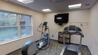 This fitness room offers a practical and well-equipped space for cardio-focused workouts. The room features a dropped ceiling with recessed fluorescent lighting panels that provide bright, even illumination throughout the space. The walls are finished in a neutral, light tone, contributing to an open and clean atmosphere.

The room is outfitted with key cardio machines, including an elliptical trainer, a treadmill, and an additional piece of cardio equipment, giving users a variety of workout options. A wall-mounted flat-screen television is positioned for easy viewing during exercise, adding an entertainment element to workout sessions. A small shelving unit sits nearby, offering convenient storage space for personal items or accessories.

Carpeted flooring with a subtle pattern adds a degree of comfort underfoot. A window along one wall allows natural light to filter into the space and provides a view of the surrounding outdoor brick exterior and greenery, creating a connection to the outside environment. The overall design is functional and straightforward, delivering the essential amenities needed for a consistent fitness routine.