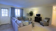 This single-floor space centers around a welcoming living room designed for comfort and functionality. The room features light-colored walls that create a bright, airy atmosphere, complemented by soft carpeted flooring that adds warmth underfoot. A large sectional sofa in a neutral gray tone serves as the focal point, adorned with decorative patterned pillows that introduce subtle texture and color to the space.

Natural light filters through a window dressed with blue-gray patterned curtains, providing both privacy and style. A wall-mounted air conditioning unit beneath the window ensures year-round comfort. On the opposite side of the room, a dark wood media console houses a flat-screen television, offering a practical entertainment setup. A tall floor lamp provides additional ambient lighting, while a lush indoor potted tree adds a natural, refreshing element to the room.

A patterned accent chair and a coffee table complete the seating arrangement, making the space ideal for both relaxing and entertaining. The overall ambiance is calm and inviting, with a cohesive neutral palette punctuated by tasteful decorative accents.