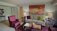 This single-floor space showcases a thoughtfully designed interior that balances comfort with contemporary style. The living area serves as the centerpiece of the home, featuring a neutral-toned sofa accented with rich burgundy and chartreuse throw pillows, creating an eye-catching contrast against the soft, light-colored walls. A large, statement artwork in warm terracotta and golden hues anchors the main wall, adding depth and a painterly focal point to the room. Two bold magenta accent chairs provide additional seating and inject a lively pop of color, complemented by a patterned area rug in deep plum and mauve tones that ties the color palette together cohesively. A sleek glass-top coffee table with a metallic frame sits at the center of the seating arrangement, topped with a decorative bowl of green apples and a blue accent piece, adding a touch of organic freshness to the space. A slender side table with a ceramic lamp provides warm ambient lighting. An archway visible in the background hints at additional rooms beyond, suggesting a functional and connected floor plan. Overall, the space exudes a vibrant, curated ambiance that is both welcoming and visually dynamic.