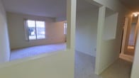This single-floor space offers a straightforward, functional layout with an open feel connecting the main living areas. The living room is carpeted and brightened by multiple windows that allow natural light to fill the room, providing views of the outdoors. Structural half-walls and archways define the transitions between spaces without fully enclosing them, contributing to an open and connected atmosphere throughout.

Adjacent to the living area, the kitchen features light-toned walls, wood-toned upper cabinetry, and a white electric range with a ventilation hood above. The kitchen transitions to a hard-surface floor, contrasting with the carpet found throughout the rest of the unit. A ceiling fan with an integrated light fixture is mounted overhead, providing both ventilation and ambient lighting to the space. A baseboard heating unit is also visible along one wall, offering supplemental climate control.

Overall, the space presents a clean, neutral palette with cream and off-white tones throughout, creating a simple and practical living environment suited for everyday use.