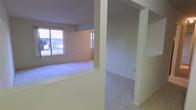 This single-floor space offers a straightforward, functional layout with an open feel connecting the main living areas. The living room is carpeted and brightened by multiple windows that allow natural light to fill the room, providing views of the outdoors. Structural half-walls and archways define the transitions between spaces without fully enclosing them, contributing to an open and connected atmosphere throughout.

Adjacent to the living area, the kitchen features light-toned walls, wood-toned upper cabinetry, and a white electric range with a ventilation hood above. The kitchen transitions to a hard-surface floor, contrasting with the carpet found throughout the rest of the unit. A ceiling fan with an integrated light fixture is mounted overhead, providing both ventilation and ambient lighting to the space. A baseboard heating unit is also visible along one wall, offering supplemental climate control.

Overall, the space presents a clean, neutral palette with cream and off-white tones throughout, creating a simple and practical living environment suited for everyday use.