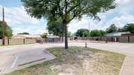 This outdoor dog park is a fully enclosed, single-level space designed to provide dogs with a safe and engaging environment for exercise and socialization. The area is surrounded by a combination of sturdy masonry walls and black metal fencing, ensuring secure containment. A prominent, mature shade tree anchors the center of the park, offering natural canopy coverage and relief from direct sunlight across much of the grounds. The surface throughout is composed of sandy, packed earth, providing a comfortable and low-maintenance ground cover suitable for active play. Several agility and enrichment features are distributed throughout the space, including a green tunnel, a tire swing, balance stepping stones, and low hurdle bars, catering to dogs of varying sizes and energy levels. A metal picnic table with bench seating is available for owners to relax while supervising their pets. A wall-mounted waste station is visible near the perimeter, supporting cleanliness and ease of use. A green low-profile platform adds an additional interactive element for smaller dogs. The park is accessed through a gated black metal entrance, marked with signage. The open-sky setting allows for ample natural light throughout the day, creating a bright and welcoming atmosphere.