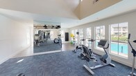 This fitness center is a thoughtfully designed, single-level space that balances architectural character with comprehensive workout functionality. The facility is divided into distinct workout zones, each tailored to a specific type of training. A standout architectural feature is the vaulted ceiling clad in natural wood planking with exposed beam detailing, lending warmth and an elevated sense of volume to the strength training area. In contrast, the cardio zone features smooth, raised ceilings with a clean, modern finish.

Large windows line the perimeter, allowing abundant natural light to fill the space and offering views of an outdoor pool area and surrounding grounds. The flooring throughout consists of durable, patterned carpet tiles in a cool blue-grey tone, providing comfort and traction underfoot.

Full-length mirrors are strategically placed along multiple walls, visually expanding the space and serving a practical purpose for form monitoring during exercise. The cardio section is well-equipped with treadmills and elliptical machines positioned to face the windows. The strength area includes cable machines, weight benches, a leg press, and a comprehensive dumbbell rack with a full range of free weights. Exercise balls and additional accessories round out the amenity offerings.