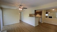 This single-floor space showcases an open-concept layout that seamlessly connects the living area to the kitchen. The living area features warm, wood-look laminate flooring that extends throughout, providing a cohesive and inviting feel. A ceiling fan with an integrated light fixture is centrally mounted on the textured ceiling, ensuring both comfort and illumination.

The kitchen is positioned at the rear of the open space and is defined by a peninsula-style breakfast bar, which serves as a natural divider while maintaining visual connectivity to the living area. Rich, dark wood cabinetry provides ample storage above, complemented by a white dishwasher and a stainless steel refrigerator. The countertops appear to feature a neutral stone-look finish. A sink is positioned along the back wall beneath upper cabinets.

A hallway visible from the living area leads further into the unit, suggesting additional private rooms beyond. The walls are painted in a soft, neutral tone throughout, contributing to a clean and understated aesthetic. White baseboards and door trim provide crisp contrast against the flooring and walls, lending the space a tidy, well-maintained appearance overall.