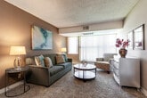 This single-floor residence offers a thoughtfully laid-out living space with a cohesive neutral color palette of warm taupes and grays throughout. The living room is carpeted and features a large window with vertical blinds that allow natural light to filter in, creating a bright and welcoming atmosphere. A vaulted ceiling detail adds an interesting architectural element to the living area. Adjacent to the living room, a separate bedroom provides a private retreat, complete with plush carpet flooring, a built-in closet with ample hanging space, and a textured ceiling. The kitchen is well-equipped with warm wood cabinetry, dark granite-style countertops, black appliances including a refrigerator, electric range, and dishwasher, all set over durable wood-look vinyl flooring. The dining area, situated near the entryway, features the same wood-look flooring and is framed by neutral walls with room for a dining table and seating. The unit includes two bathrooms: one with a single-basin vanity, wood cabinetry, and a walk-in shower, and another with a clean white soaking tub and tiled surround. Each room is finished with textured ceilings and clean white trim, contributing to a consistent and comfortable aesthetic throughout.