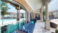 This covered outdoor lounge offers a welcoming retreat that seamlessly blends classic architectural design with comfortable, resort-style amenities. The exterior facade features warm red brick accented by elegant white columns that support the covered overhang, creating a shaded corridor perfect for year-round use. Large arched glass doors and windows line the interior wall, allowing abundant natural light to filter through while providing direct visual access to the sparkling pool area just beyond. The flooring consists of a neutral stone tile laid in a geometric pattern, adding a refined, polished aesthetic to the space. Plush, navy blue striped sofas with vibrant teal accent pillows are arranged throughout the lounge, offering ample seating for socializing or unwinding. The open-air design allows for natural ventilation while the overhead covering ensures shade and shelter. Lush greenery is visible at the perimeter, softening the architectural elements and enhancing the outdoor ambiance. Overall, this lounge delivers an inviting, upscale atmosphere with a strong connection to the surrounding outdoor amenities.