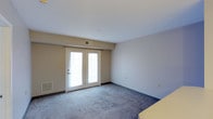 This single-floor space offers a clean, straightforward layout designed for comfortable living. The main living area is finished with wall-to-wall carpet in a neutral blue-grey tone, providing a soft and practical underfoot surface. Smooth, white ceilings give the room an open, airy feel, while the neutral beige and grey wall tones create a calm, versatile backdrop suitable for a variety of interior styles.

A standout feature of the space is the set of white-framed French doors, which serve as both a functional entryway and a significant source of natural light. The doors allow daylight to filter throughout the room, enhancing the overall brightness of the interior. A wall-mounted HVAC vent is positioned above the French doors, and a floor-level heating unit is also visible nearby, ensuring year-round climate comfort.

The room includes multiple electrical outlets along the walls, offering practical placement for furniture and appliances. Dark baseboards provide a defined contrast against the lighter wall tones, adding a subtle architectural detail throughout the space. Overall, the interior presents a functional, move-in-ready environment with a neutral palette that offers flexibility for personalization.