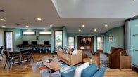 This single-floor community clubhouse is thoughtfully designed to serve as a multifunctional gathering space. The lounge area features mid-century modern furnishings, including a tan leather sofa, blue accent chairs, and a colorful area rug layered over warm hardwood flooring. A wall-mounted flat-screen TV is built into a wooden media unit, while recessed lighting and a tray ceiling add architectural interest overhead. Teal-painted walls provide a vibrant, welcoming backdrop throughout the space, complemented by natural light streaming through multiple windows. A dedicated computer workstation area offers a row of desktops along a wooden counter, ideal for productivity. The dining zone showcases a whitewashed exposed brick accent wall adorned with framed artwork, paired with dark wood tables and industrial-style metal chairs. A translucent glass partition wall separates functional zones while maintaining visual openness. The kitchenette is well-appointed with granite countertops, dark gray cabinetry, stainless steel appliances including a microwave and refrigerator, and direct access to an outdoor patio through glass-paneled doors. Pendant lighting and a water dispenser complete the functional amenities, making this a versatile and stylish community hub.