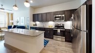 This single-floor residence offers a thoughtfully designed open-concept layout that seamlessly connects the kitchen, dining, and living areas. The kitchen serves as the centerpiece, showcasing rich dark cabinetry with brushed nickel hardware, granite countertops, and a full suite of stainless steel appliances including a range, microwave, dishwasher, and refrigerator. A generously sized kitchen island with matching granite provides additional workspace and casual seating, while two globe pendant lights suspended overhead add a contemporary touch. Light wood-tone flooring runs throughout the main living areas, enhancing the bright, neutral color palette.

The living room is well-proportioned with multiple windows dressed in layered blinds and curtains, allowing abundant natural light. A ceiling fan keeps the space comfortable.

The unit includes two bedrooms, each featuring windows with natural light, ceiling fans, and wood-tone flooring. The primary bathroom is appointed with a dark vanity, granite countertop, undermount sink, and large mirror. An adjacent walk-in closet with wire shelving provides generous storage. Overall, the space offers a clean, modern aesthetic with upscale finishes throughout.